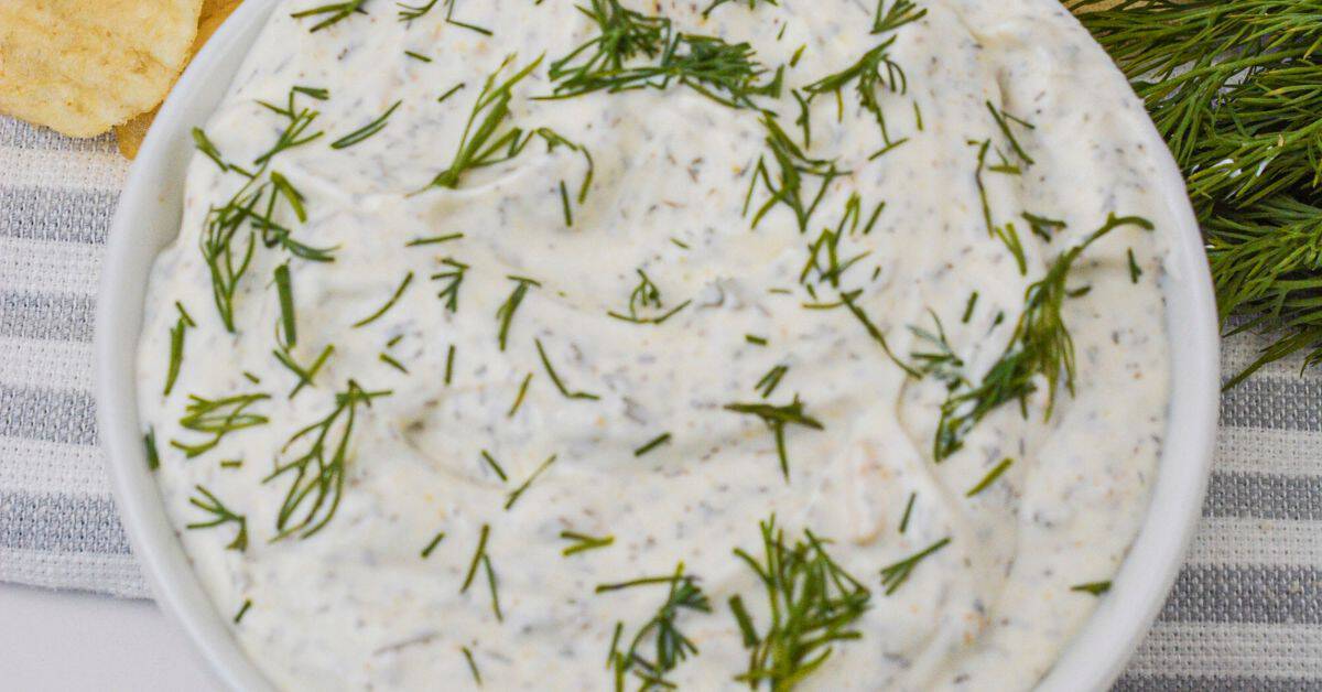 A bowl of creamy Dill Dip topped with chopped fresh dill sits on a striped cloth, surrounded by potato chips and sprigs of dill.