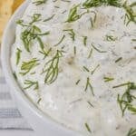 A white bowl filled with creamy dill dip garnished with fresh dill, with chips visible in the background.