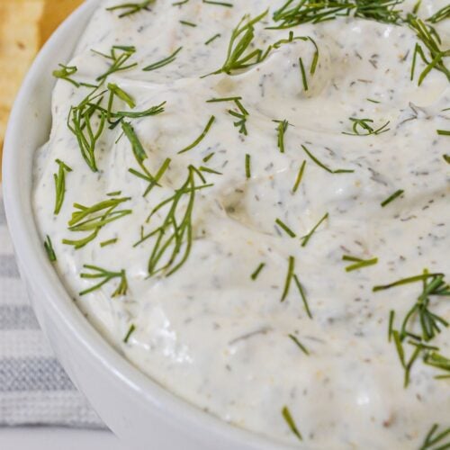 A white bowl filled with creamy dill dip garnished with fresh dill, with chips visible in the background.