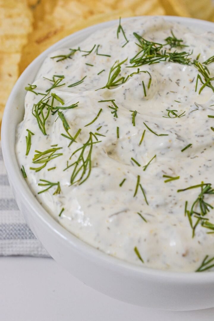 A white bowl filled with creamy dill dip garnished with fresh dill, with chips visible in the background.