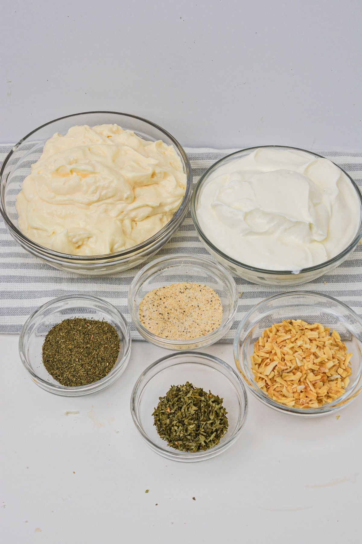 Six glass bowls containing ingredients —mayonnaise, sour cream, dried parsley, dried chives, dried dill, and dried onion—are arranged on a striped cloth.