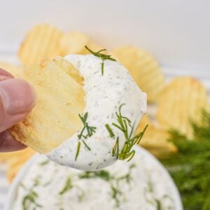 A hand holds a ridged potato chip dipped in creamy dill dip, garnished with fresh dill, with more chips and sprigs of dill in the background.