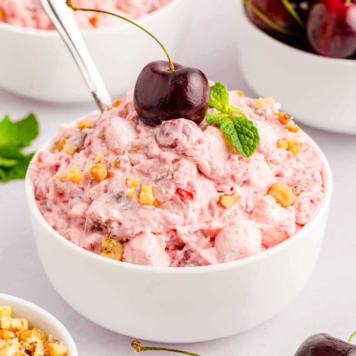 A white bowl filled with pink cherry fluff salad, topped with a whole cherry, chopped nuts, and a mint sprig; a spoon is in the bowl. It’s a Keeper dessert that looks as good as it tastes!.