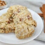 Three glazed Cinnamon Scones with visible pecan pieces sit on a white plate. In the background are two cinnamon sticks and a small bowl of chopped pecans.