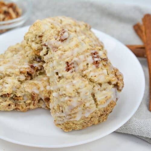 Three glazed Cinnamon Scones with visible pecan pieces sit on a white plate. In the background are two cinnamon sticks and a small bowl of chopped pecans.