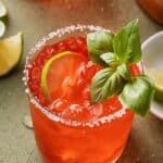 A red summer cocktail garnished with a lime slice and fresh basil in a salt-rimmed glass, with lime wedges nearby. Text reads: "10 Refreshing Summer Cocktails—perfect summer drinks to cool you off.