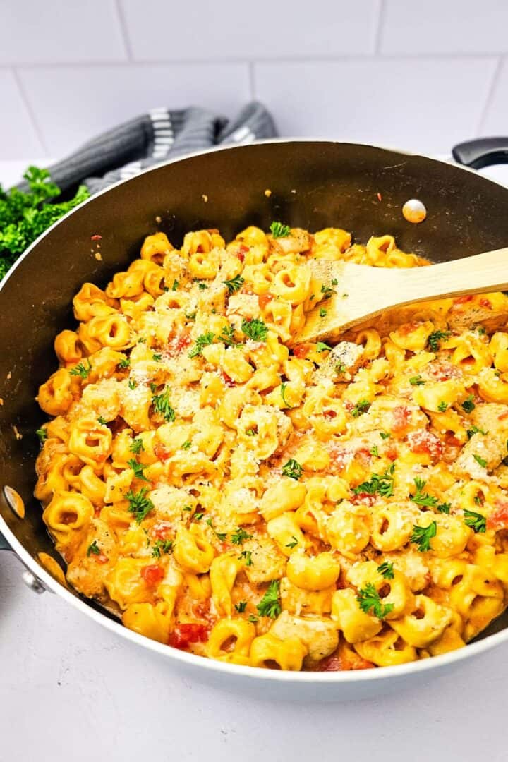 A skillet filled with creamy chicken tortellini pasta, mixed with sauce, herbs, and grated cheese, with a wooden spoon resting on the side—a comforting skillet recipe perfect for any night of the week.