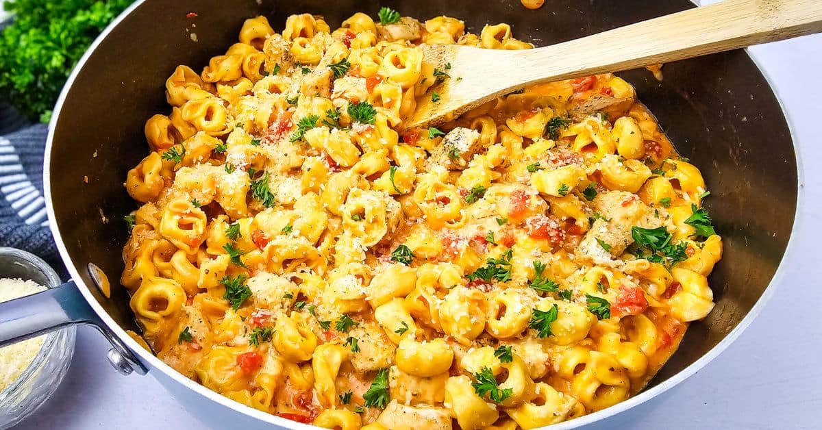 A pan of creamy chicken tortellini pasta with tomatoes, sprinkled with chopped parsley and grated cheese, stirred with a wooden spoon—this easy skillet recipe is perfect for a comforting weeknight meal.