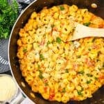 A pan filled with creamy pasta, topped with parsley and grated cheese, with a wooden spoon resting inside. This easy skillet recipe is completed by a bowl of grated cheese and a kitchen towel nearby.