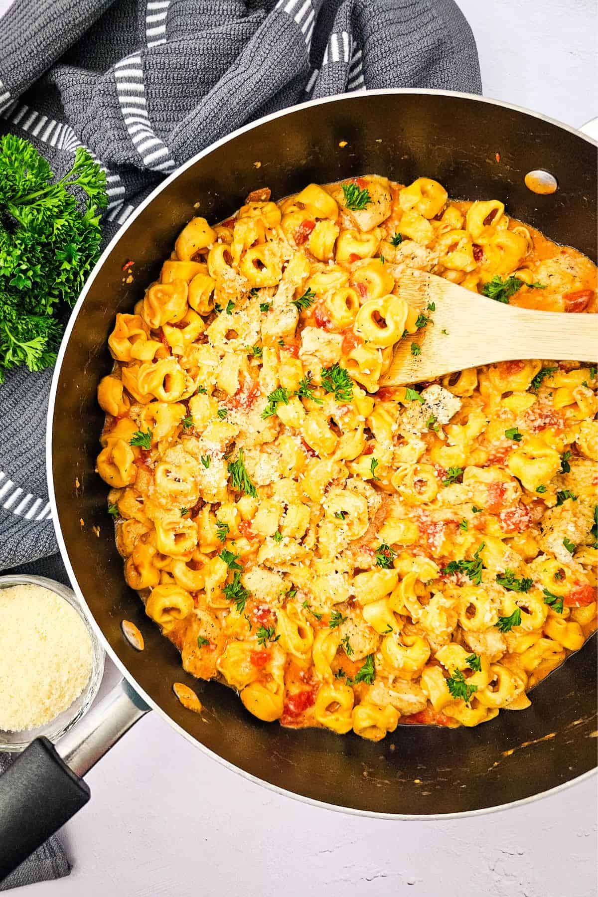 A pan filled with creamy  pasta, topped with parsley and grated cheese, with a wooden spoon resting inside. This easy skillet recipe is completed by a bowl of grated cheese and a kitchen towel nearby.