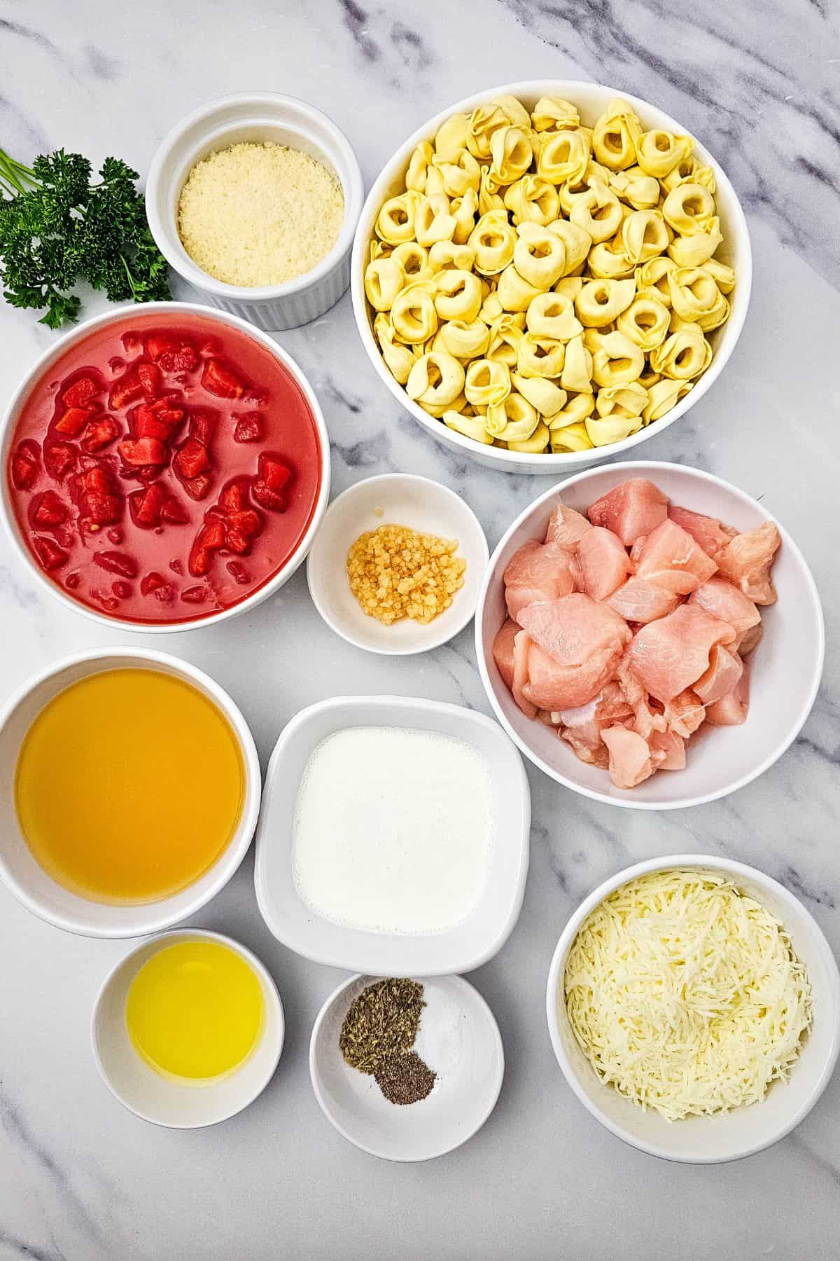 Various ingredients in bowls on a marble surface, including chicken tortellini, diced tomatoes, chicken pieces, cheeses, garlic, broth, cream, oil, spices, and parsley.