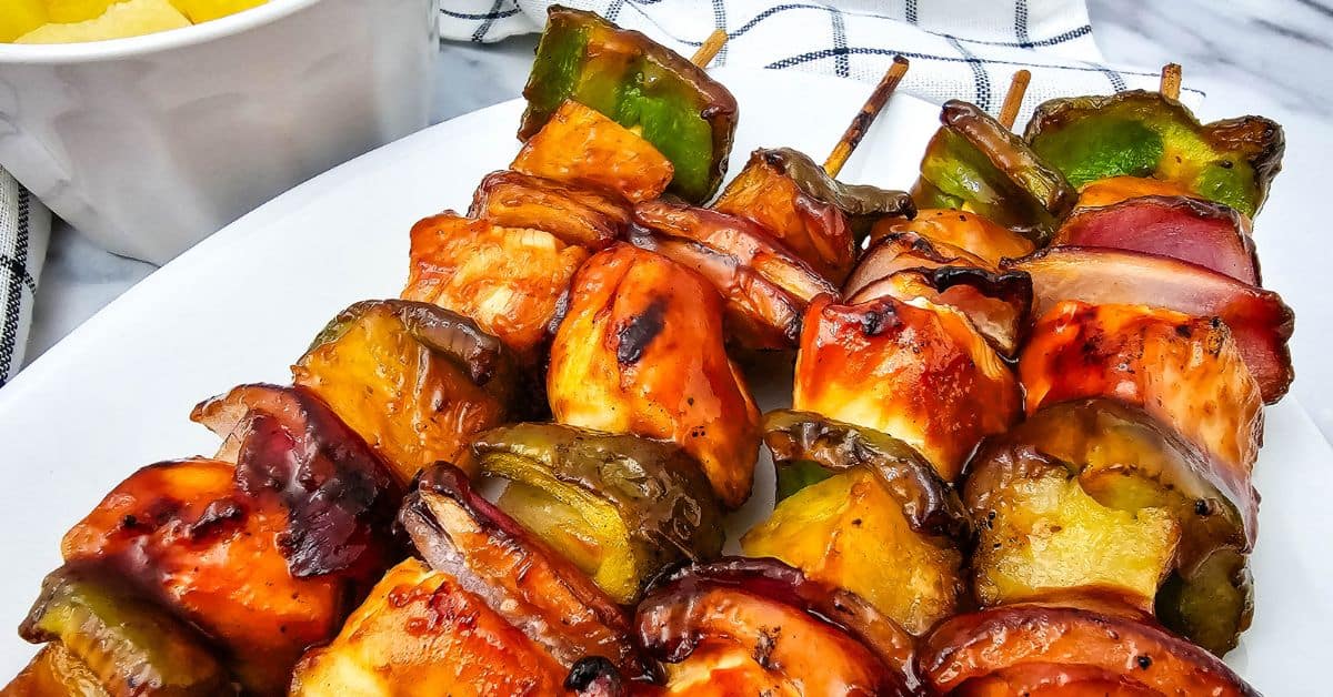Grilled BBQ Chicken skewers with chunks of chicken, green bell pepper, red onion, and pineapple arranged on a white plate.