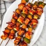 Three grilled chicken kabobs with bell peppers, red onion, and pineapple are served on a white oval plate; a bowl of pineapple chunks sits beside the plate on a marble surface.