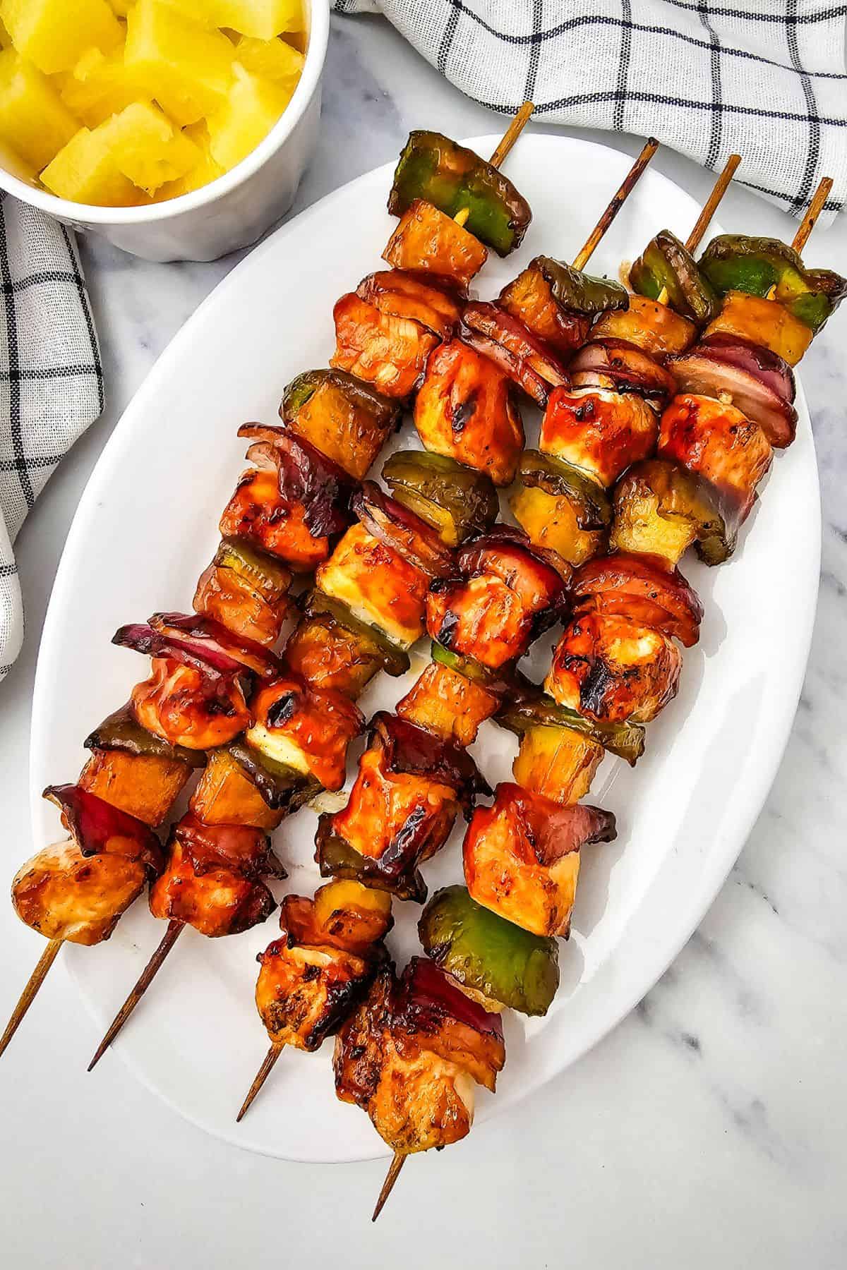 Three skewers with bell peppers, red onion, and pineapple are served on a white oval plate; a bowl of pineapple chunks sits beside the plate on a marble surface.