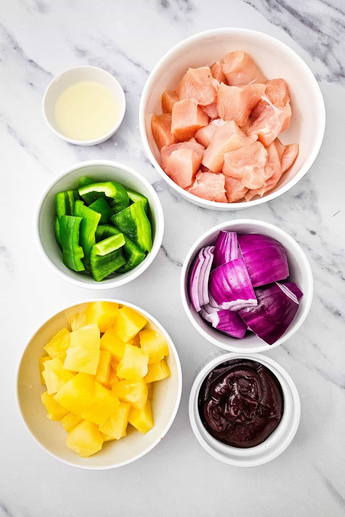 Six bowls on a marble surface contain the ingredients: raw chicken, green bell pepper, red onion, pineapple chunks, a dark BBQ sauce, and a small amount of liquid—each arranged neatly.
