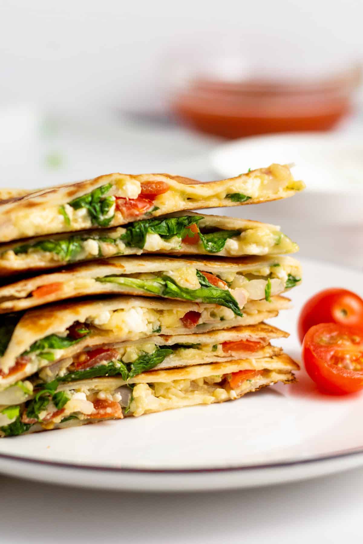 Stack of sliced quesadillas filled with eggs, cheese, greens, and tomatoes on a white plate, with cherry tomato halves beside them—a perfect addition to your collection of easy Girl Dinners or Lazy Meals.