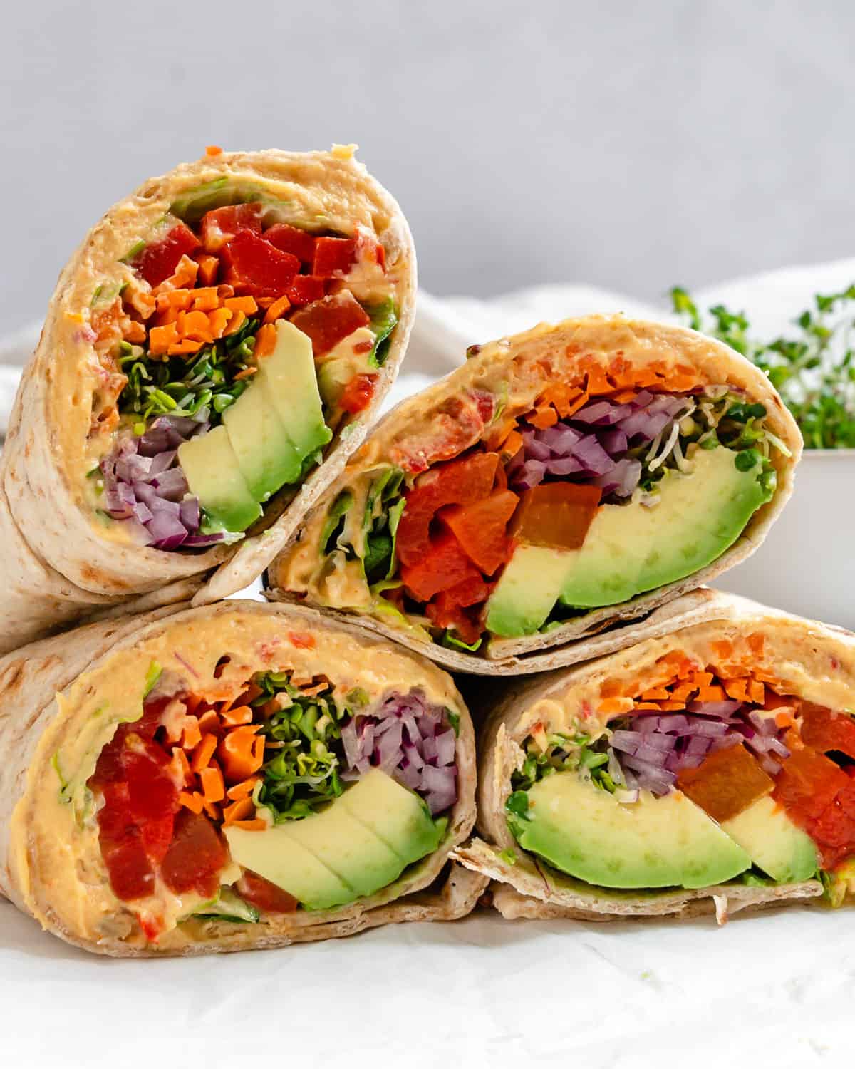 Four vegetable wraps cut in half, showing layers of avocado, shredded carrot, red onion, greens, and hummus inside whole wheat tortillas, stacked on a white surface—perfect for those lazy meals or quick Girl Dinners when you want something fresh and satisfying without spending hours in the kitchen.