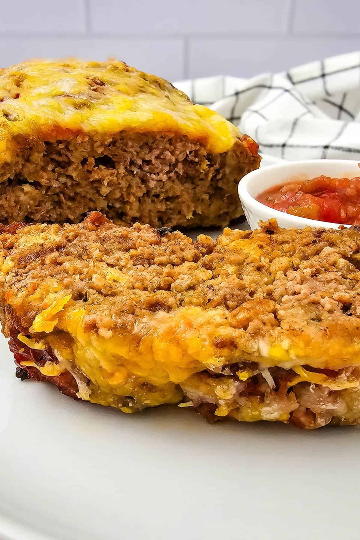 Sliced salsa meatloaf topped with melted cheese, served with a small bowl of red sauce.