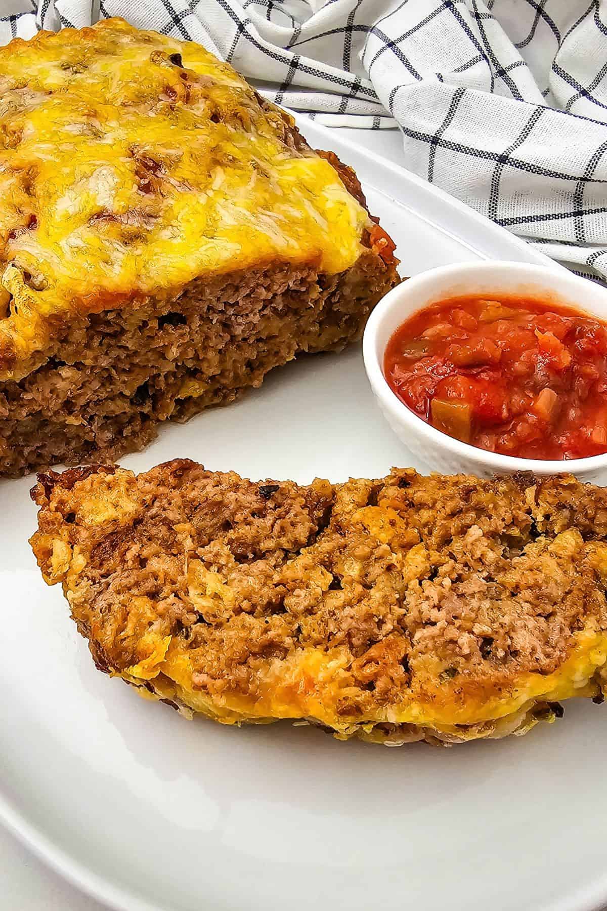 A loaf of salsa meatloaf topped with melted cheese, a sliced serving, and a small dish of salsa on a white platter.