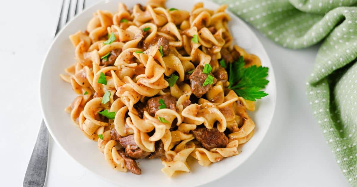 Beef stroganoff with egg noodles on a white plate, garnished with parsley; a classic dish of slow cooker beef.