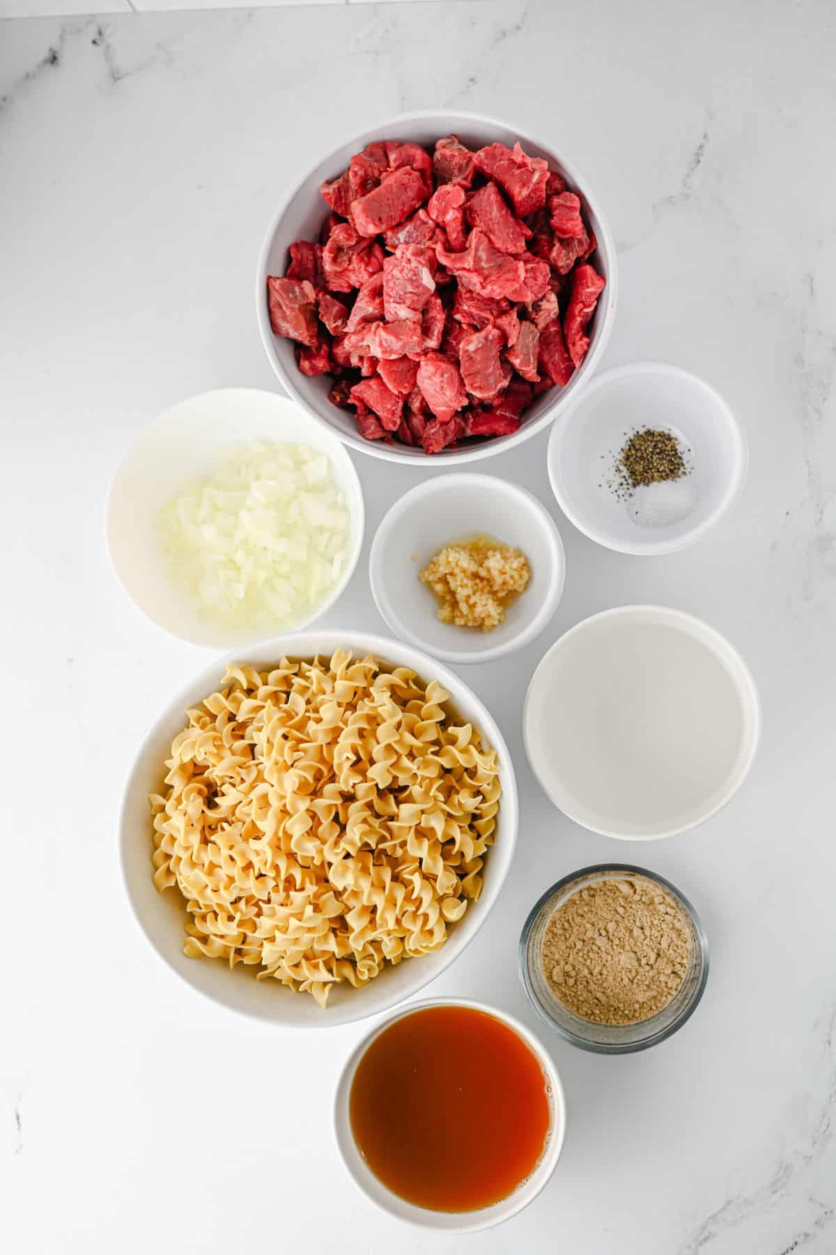 Overhead view of ingredients for crock pot beef stroganoff: cubed beef, egg noodles, onion, garlic, broth, seasonings, and cream sauce.