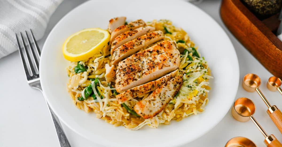 Sliced lemon chicken served over orzo pasta with spinach, topped with a fresh lemon wedge.