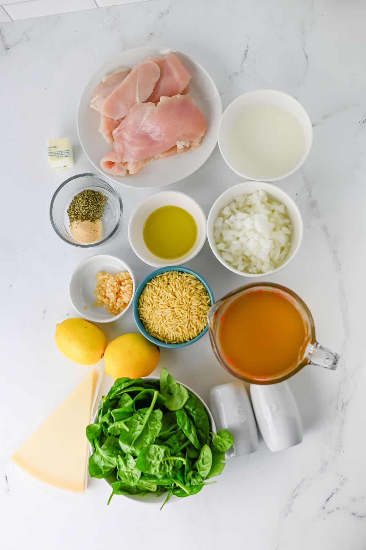 Ingredients for a lemon chicken recipe arranged on a white surface, including raw chicken breasts, orzo, spinach, lemons, broth, milk, oil, butter, diced onion, garlic, spices, and Parmesan cheese.