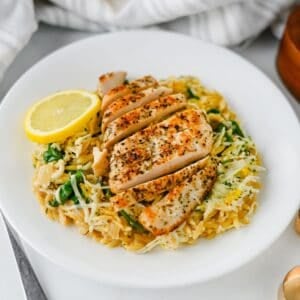 Sliced, seasoned lemon chicken served over orzo pasta with spinach and cheese, garnished with a lemon slice on a white plate.
